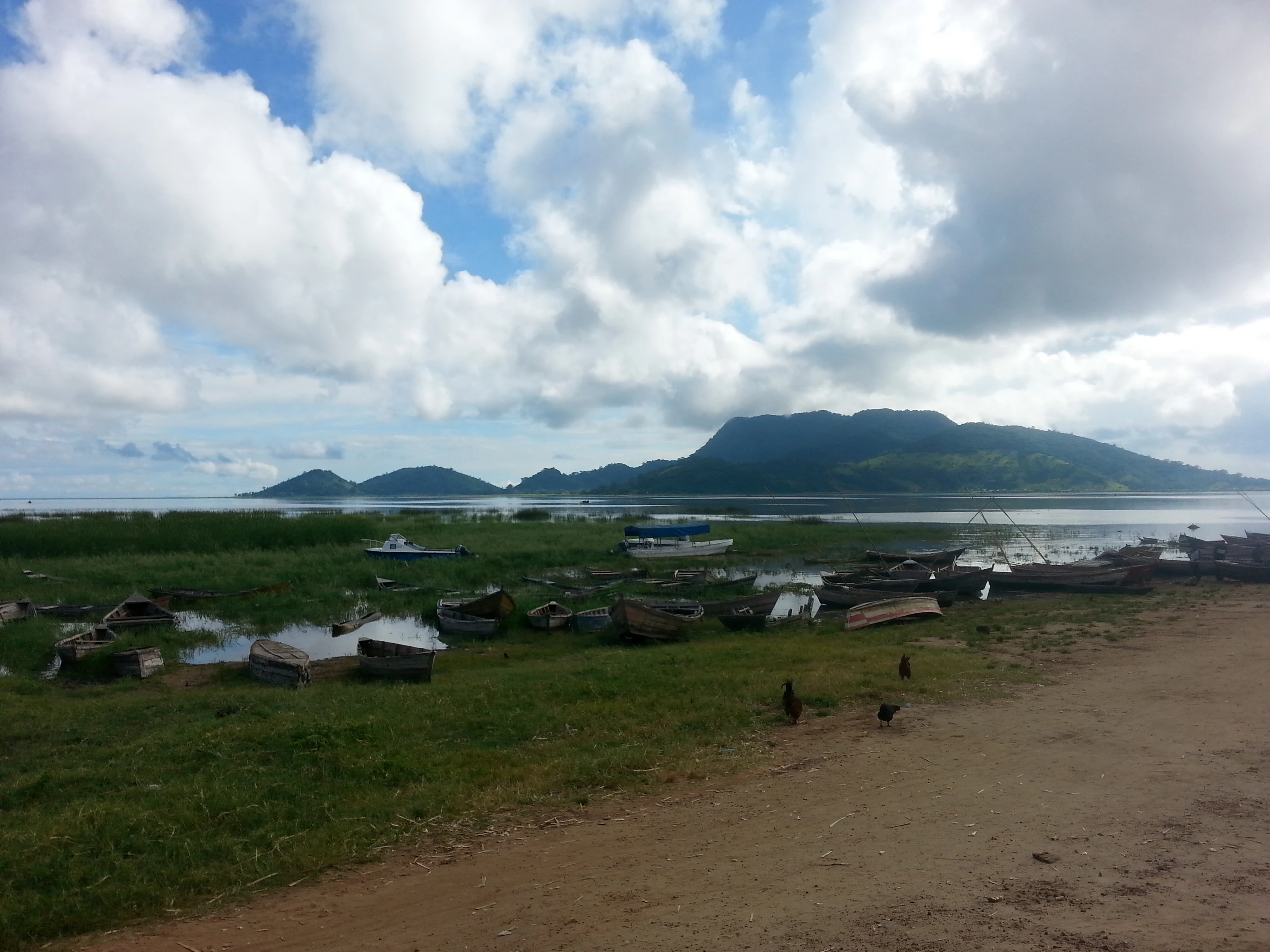 A wide view of the grassy shore of Lake Chilwa with small wooden fishing boats moored in shallow water, and a large hill, likely Chisi Island, rising in the background under a cloudy sky.