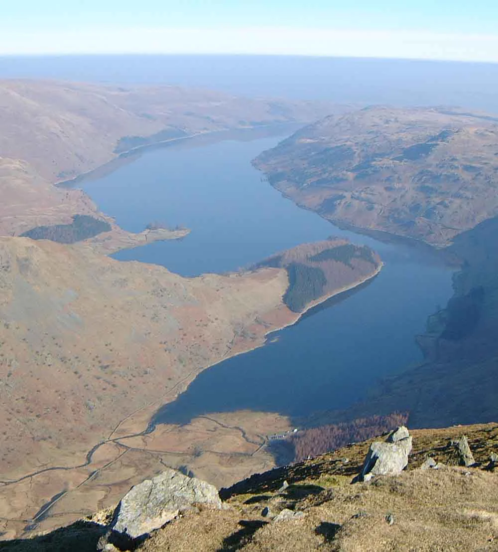 Wide aerial view of a large lake surrounded by rolling hills and mountains, with rocky outcrops in the foreground