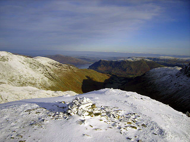 Snow-covered mountain summit with a stone cairn, showing a valley with snow-covered mountains and a body of water in the distance