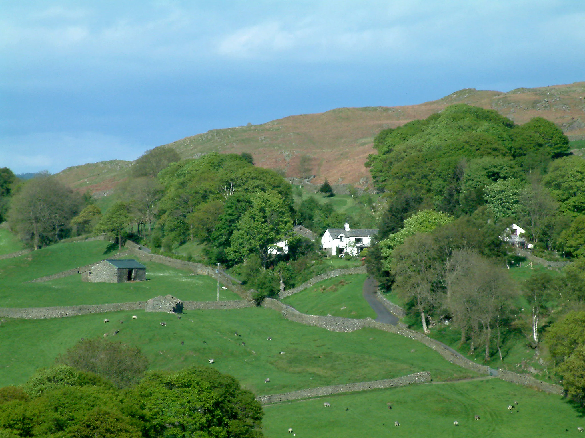 Green fields with stone walls, stone barn, white houses, trees, and a hill under a partly cloudy sky