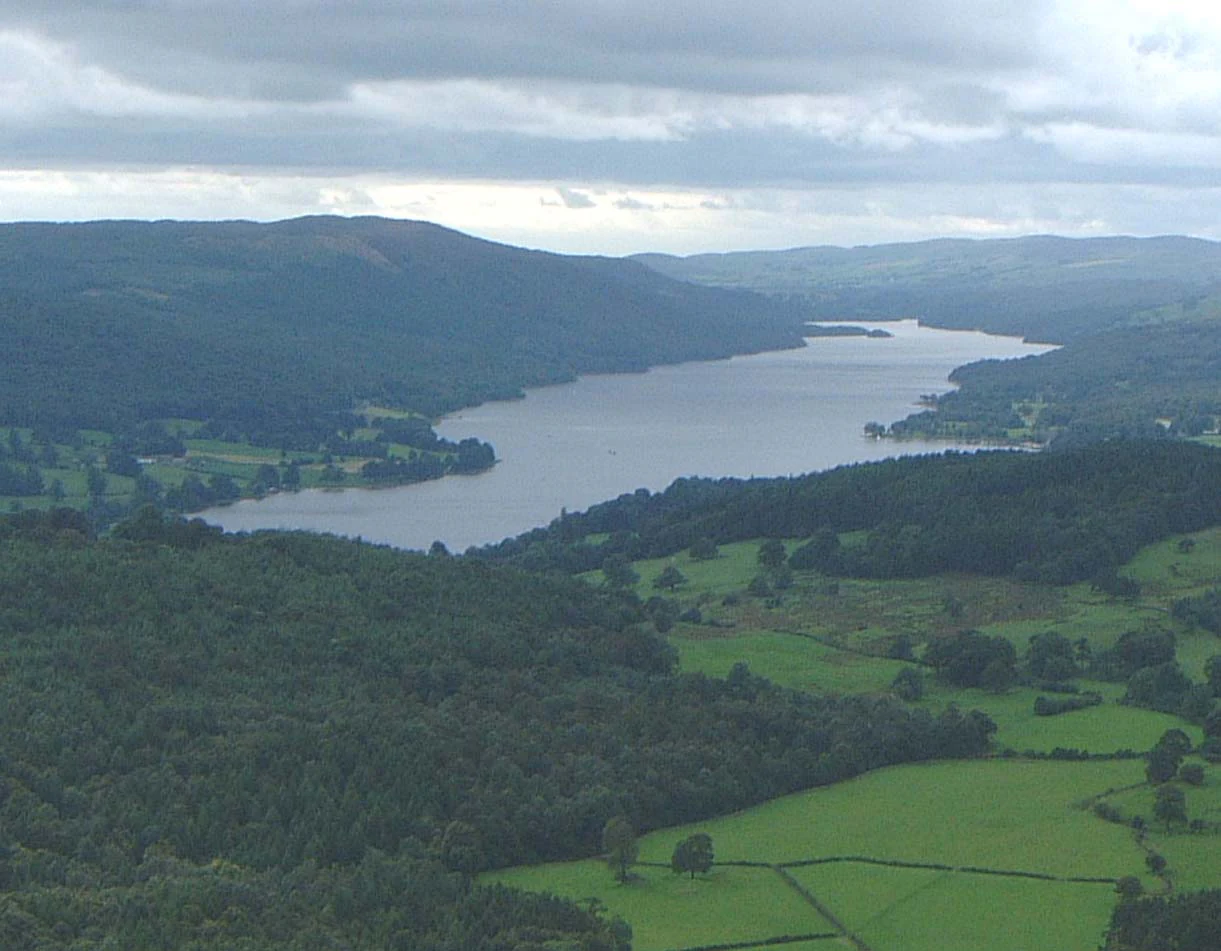 Panoramic view of a large lake bordered by rolling green hills and dense forests under a partly cloudy sky.