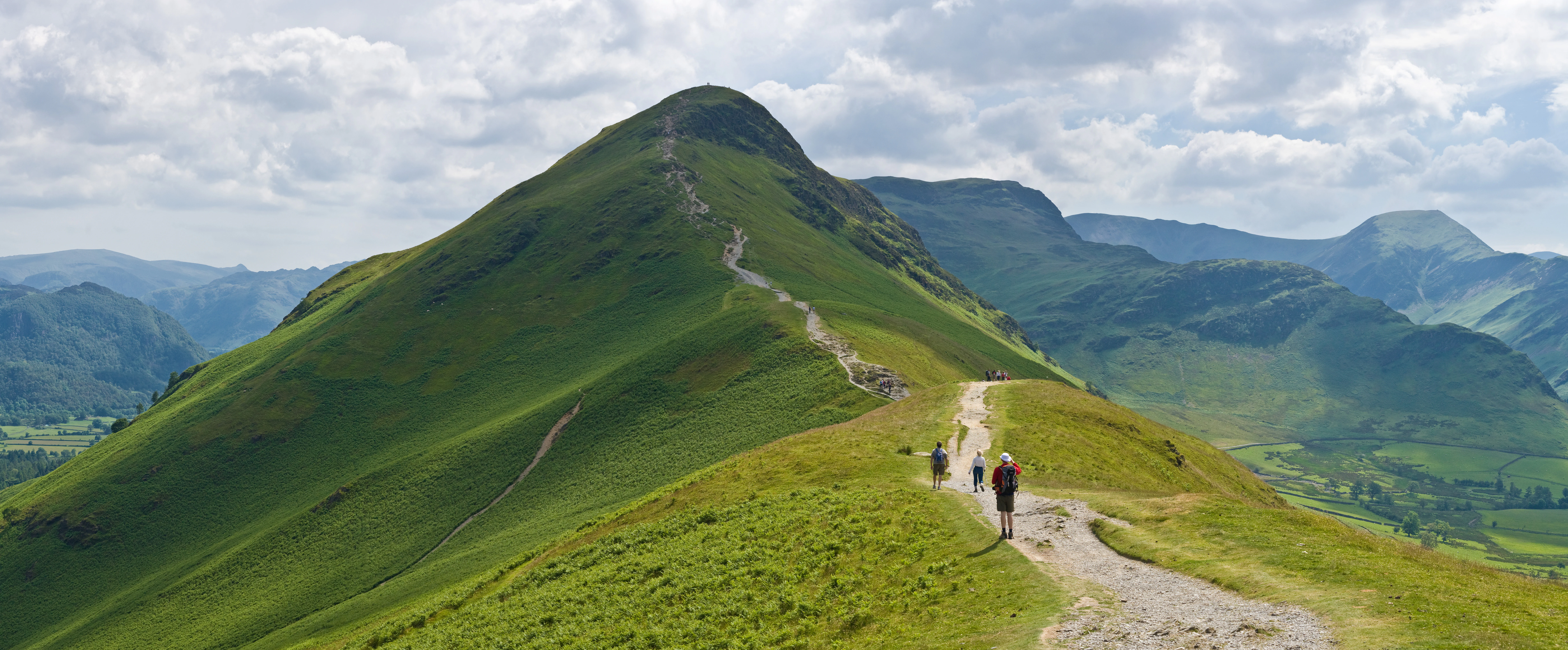A scenic view of Catbells mountain in Lake District National Park, showing a trail with hikers ascending the green slope under a partly cloudy sky