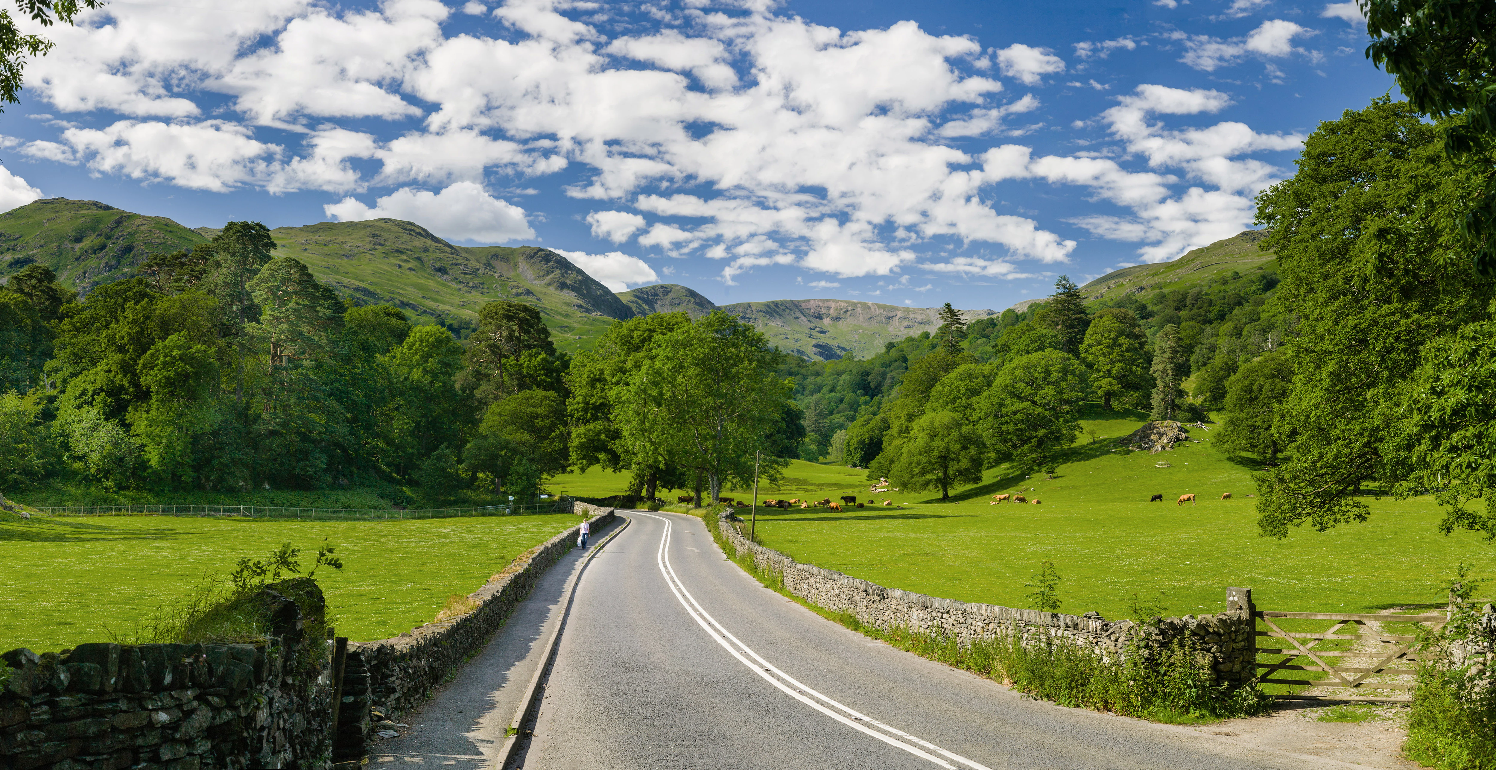 Paved road with white lines flanked by stone walls, green fields, and trees under a blue sky with scattered clouds