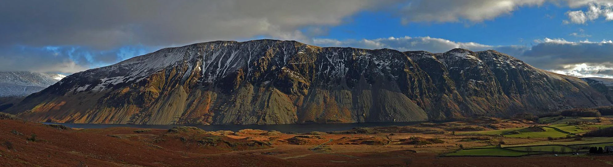 A panoramic view of Wasdale Screes showing rocky mountain slopes with patches of snow, a valley with fields, and a partly cloudy sky