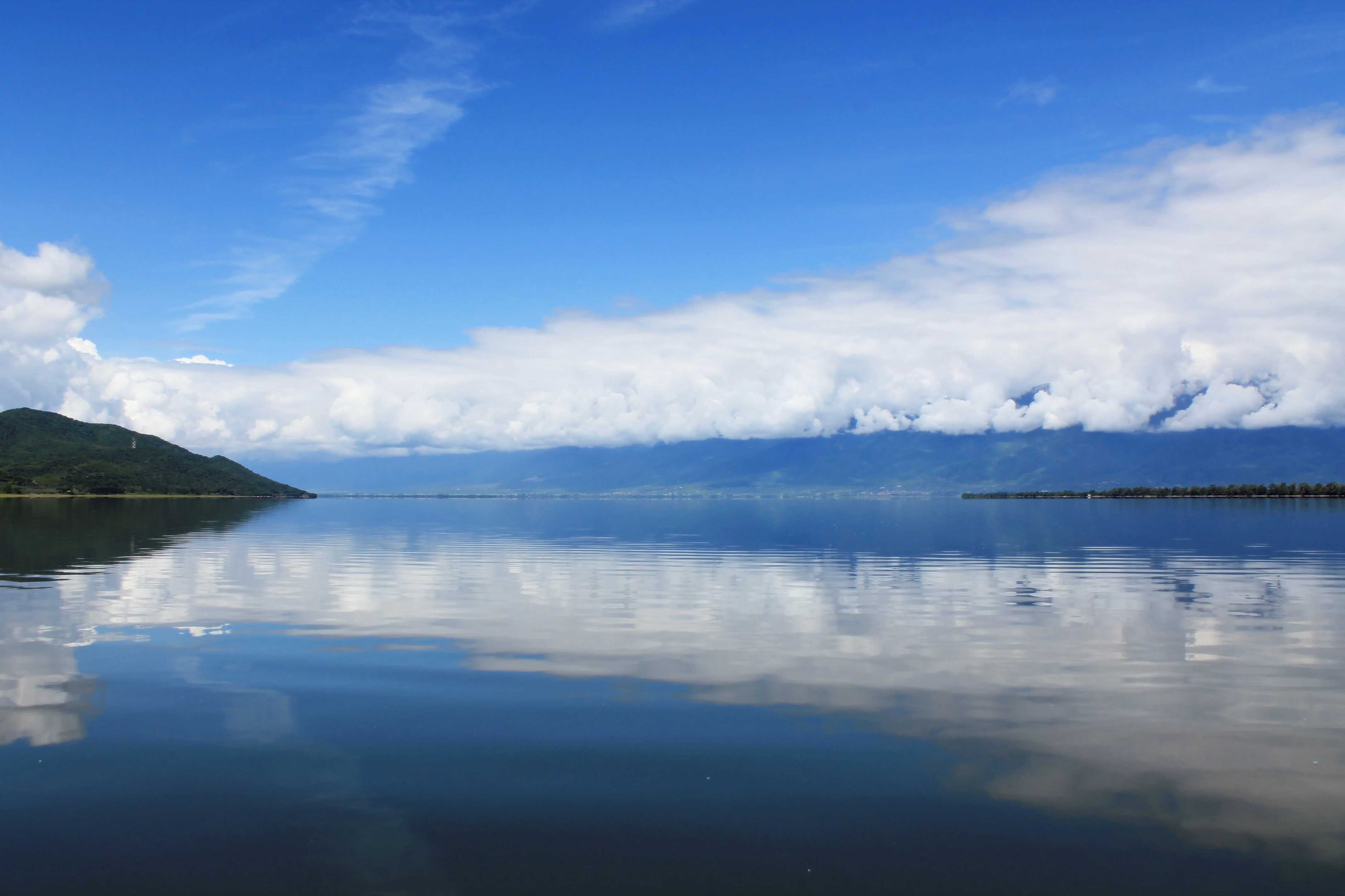 A wide landscape view of a calm lake reflecting a blue sky with large white clouds, with a green hill on the left and a line of trees on the distant horizon.