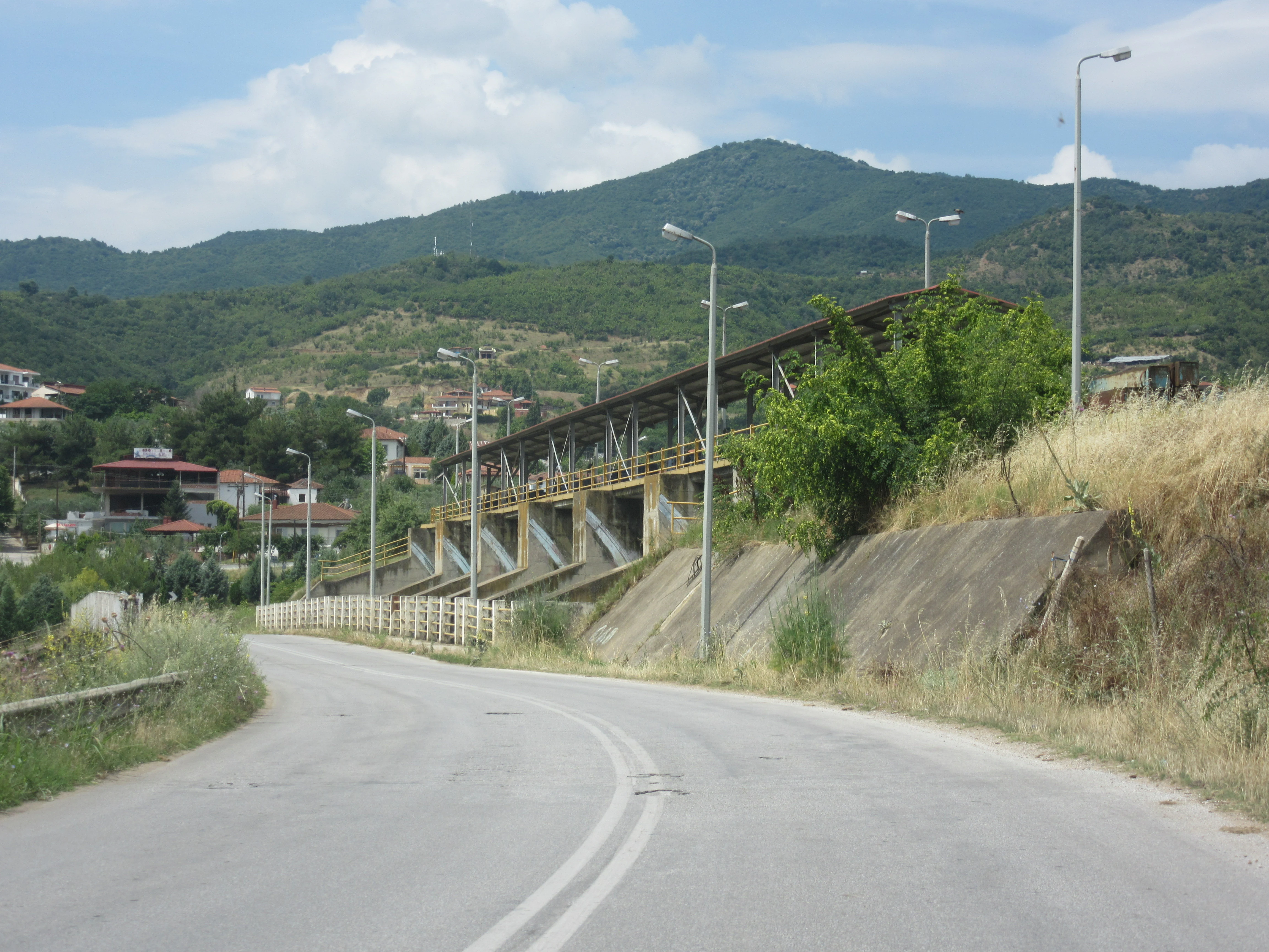 A paved road curves towards a large concrete dam with multiple spillway gates, set against a backdrop of green hills and a blue sky with clouds.