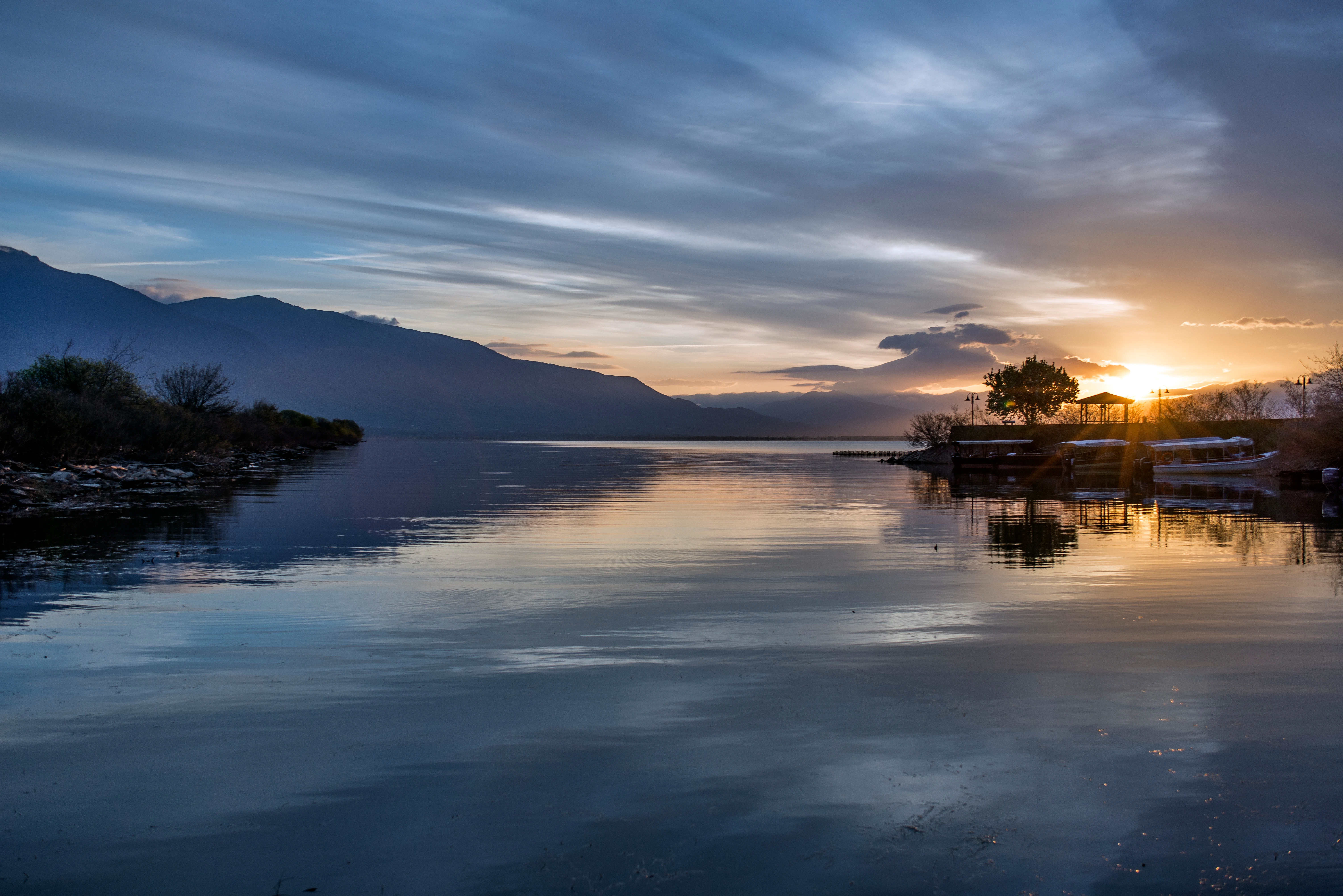 A wide landscape shot of a lake at sunset, featuring calm water reflecting the sky, mountains in the distance on the left, and a small port with trees and boats silhouetted against the sun on the right.