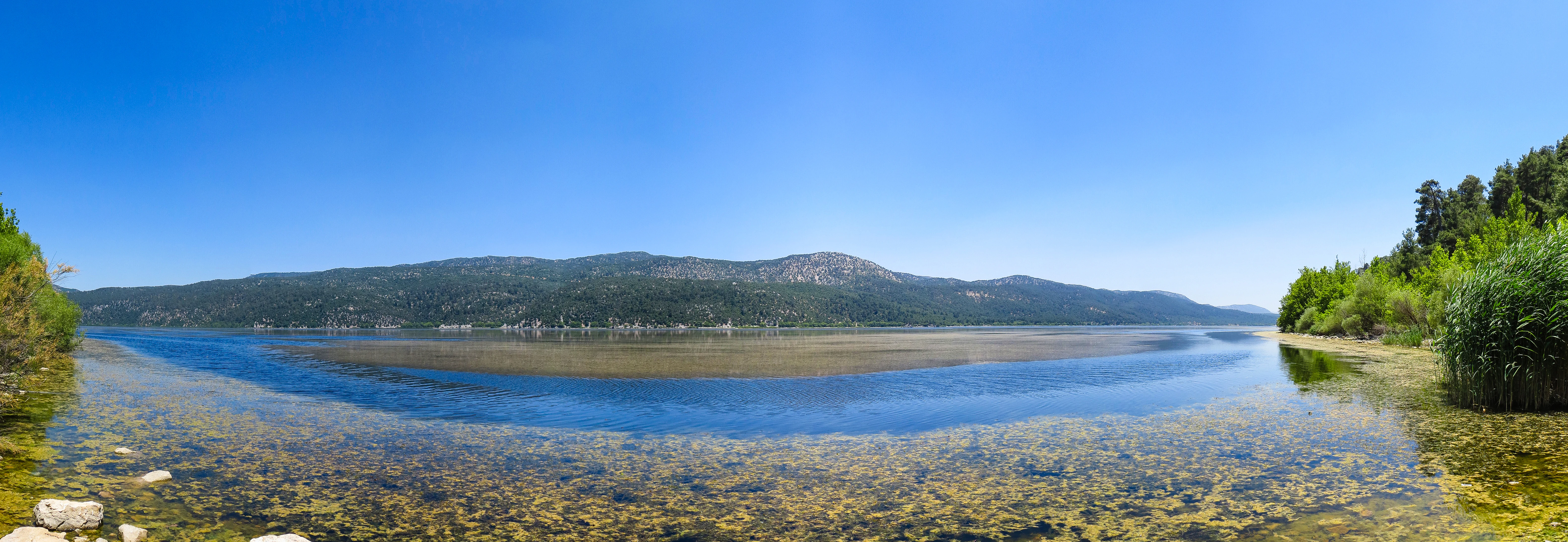 A panoramic view of Lake Kovada showing clear shallow water, rocky shore, and mountainous backdrop under a clear blue sky