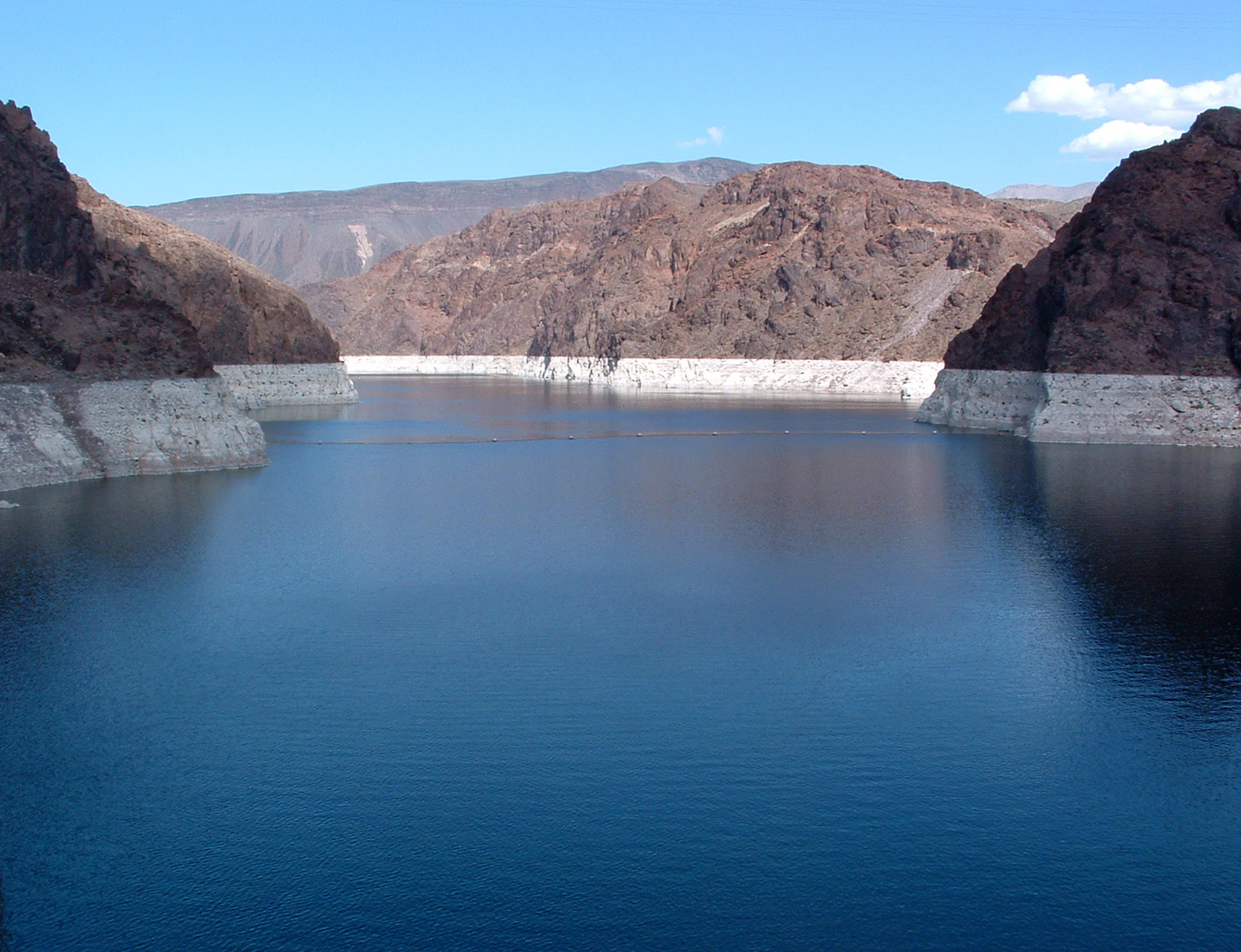 A wide landscape photograph showing deep blue water of Lake Mead framed by dark, rocky canyon walls, with a distinct light colored 'bathtub ring' mineral deposit visible along the shoreline.