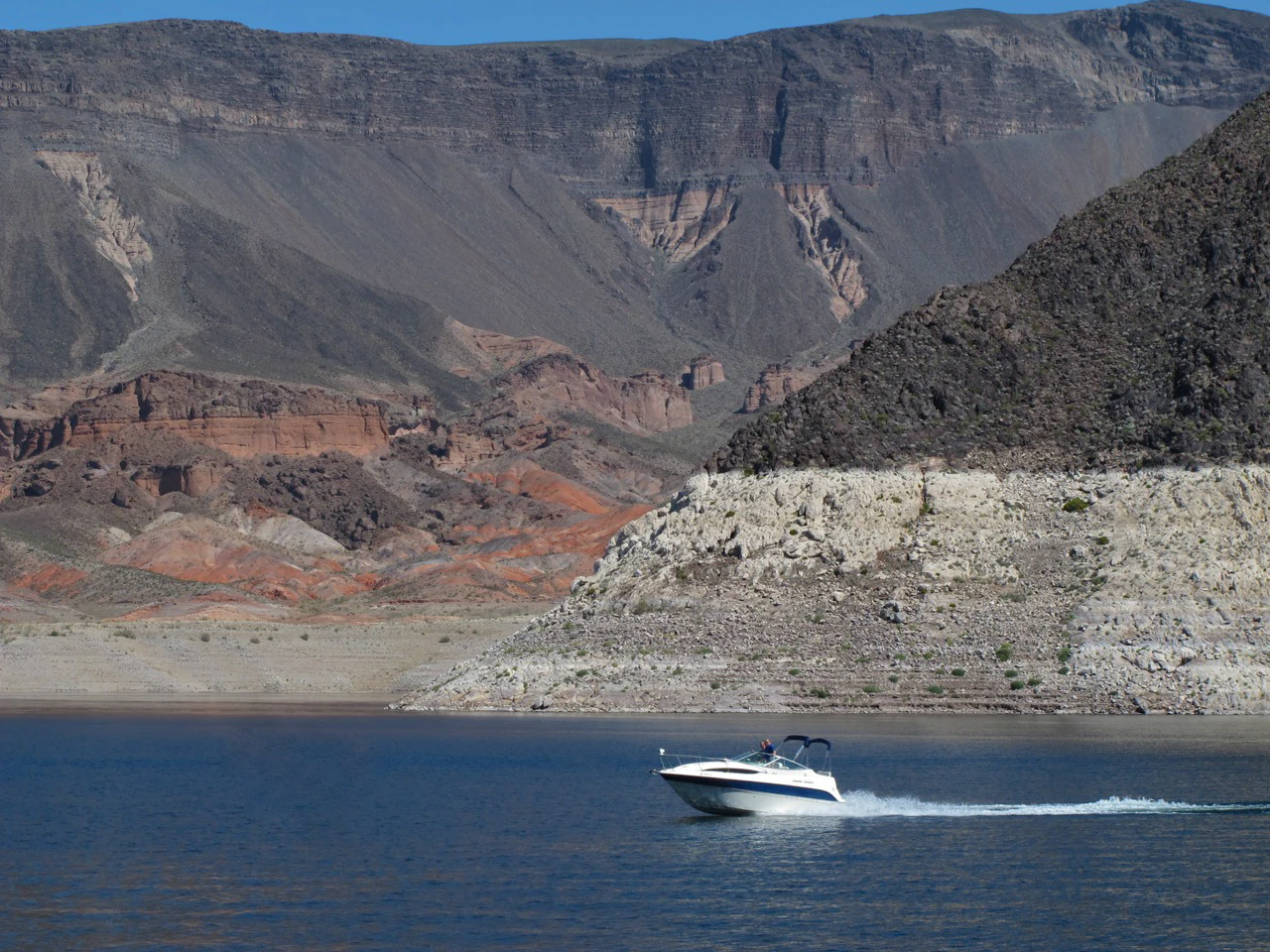 A small white motorboat creating a wake while traveling across dark blue water in front of barren, rocky desert cliffs.