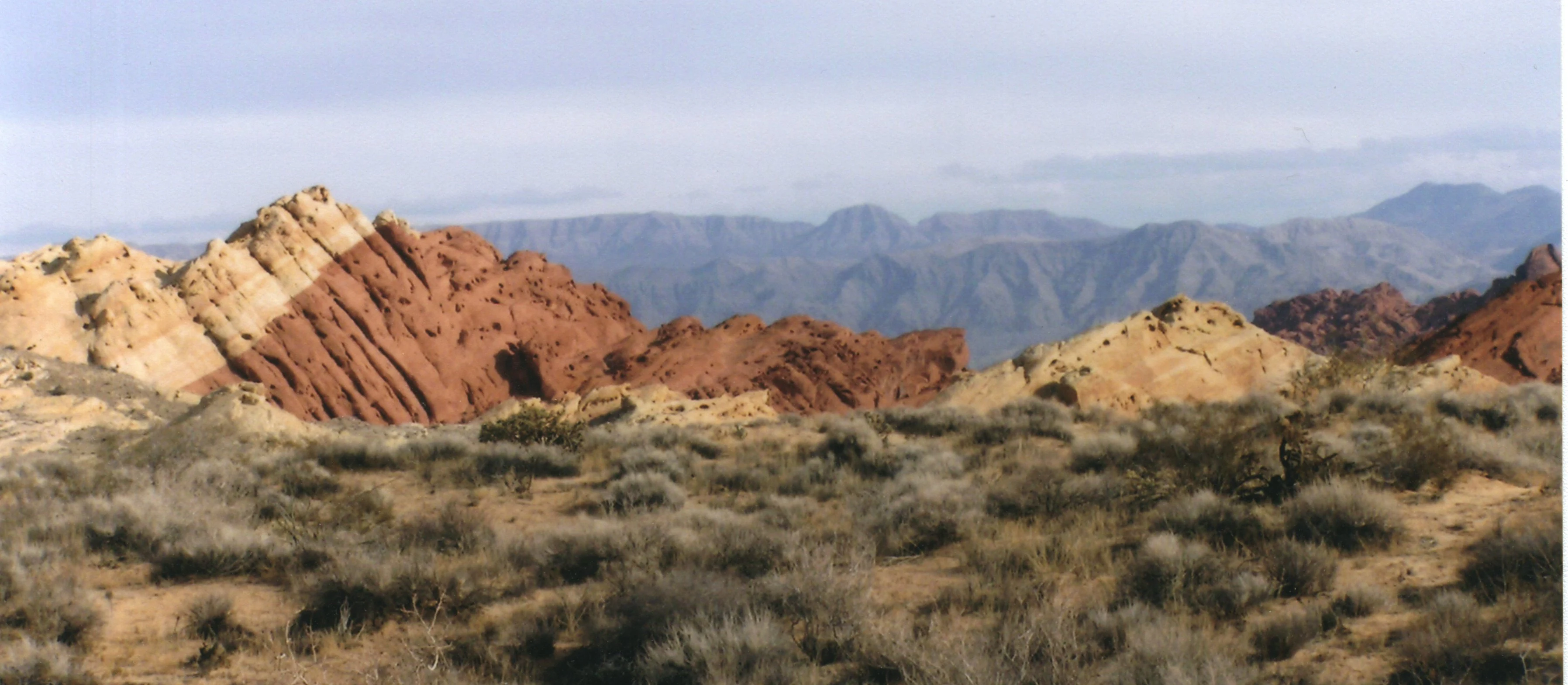 A landscape view showing prominent red and tan sandstone rock formations set against a desert scrub foreground and distant blue-toned mountains.