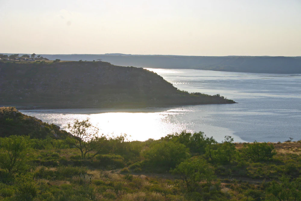 A wide landscape view of a reservoir at sunset with high bluffs rising from the water and scrubby vegetation in the foreground.