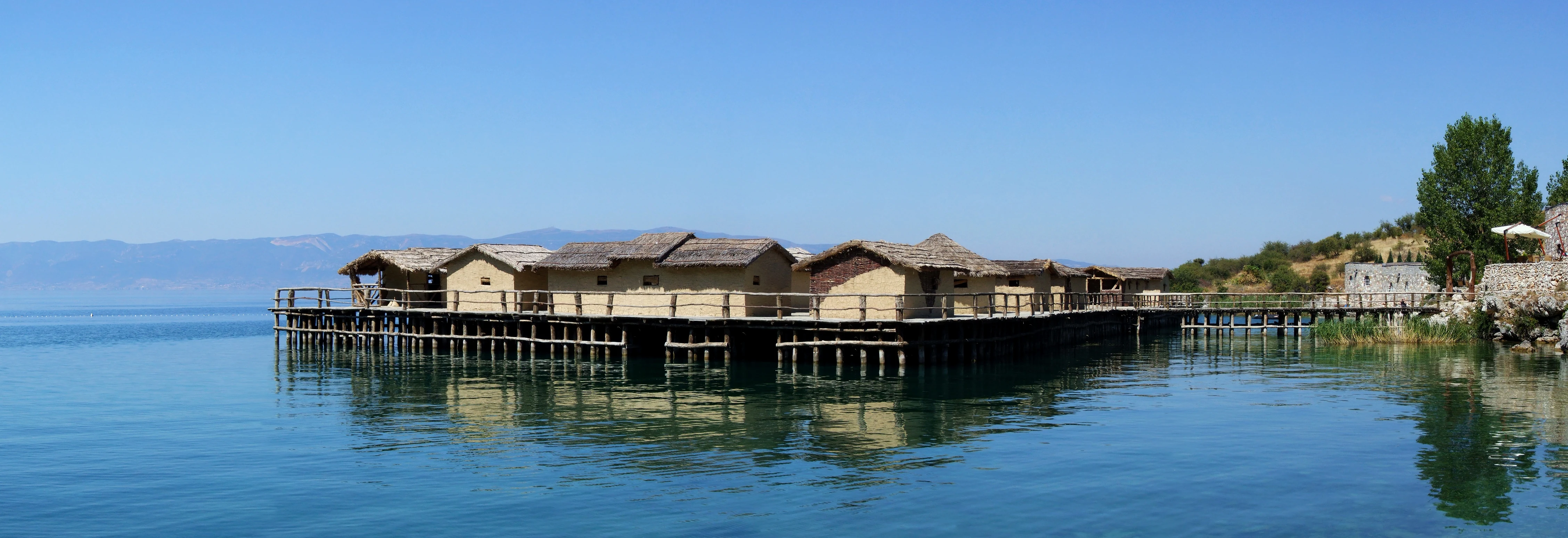 A reconstructed prehistoric pile dwelling village built on wooden stilts over the clear, calm blue water of Lake Ohrid.
