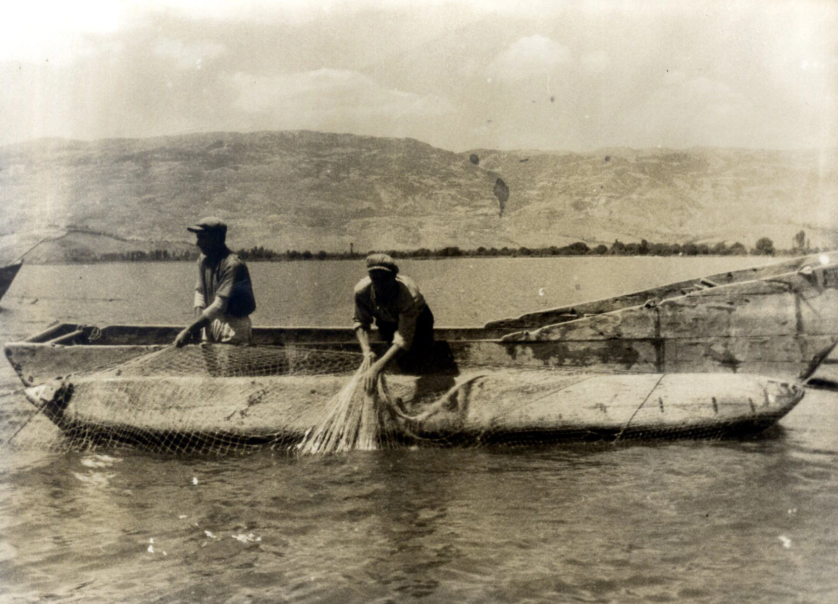 A sepia-toned vintage photograph showing two men in a flat-bottomed wooden boat on Lake Ohrid, pulling in fishing nets. A shoreline and hills are visible in the background.