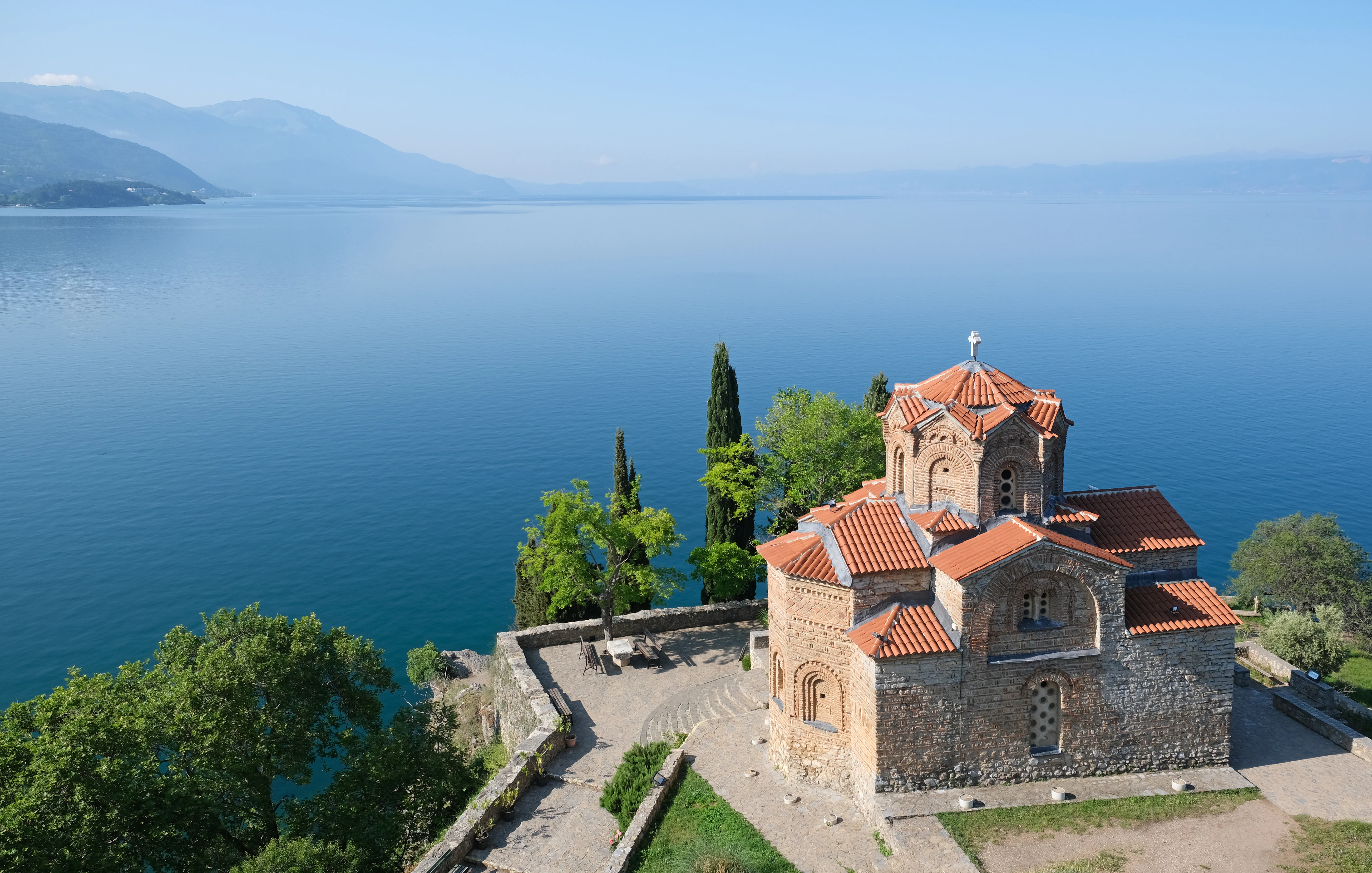 An elevated view of the stone-built Church of St. John at Kaneo with a terracotta roof, situated on a cliffside overlooking the expansive blue expanse of Lake Ohrid.