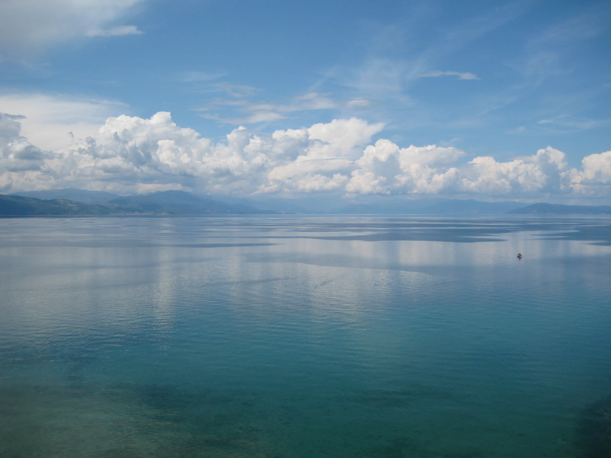 A wide, elevated view of a calm, expansive lake with clear blue water and distant hills under a partly cloudy sky.