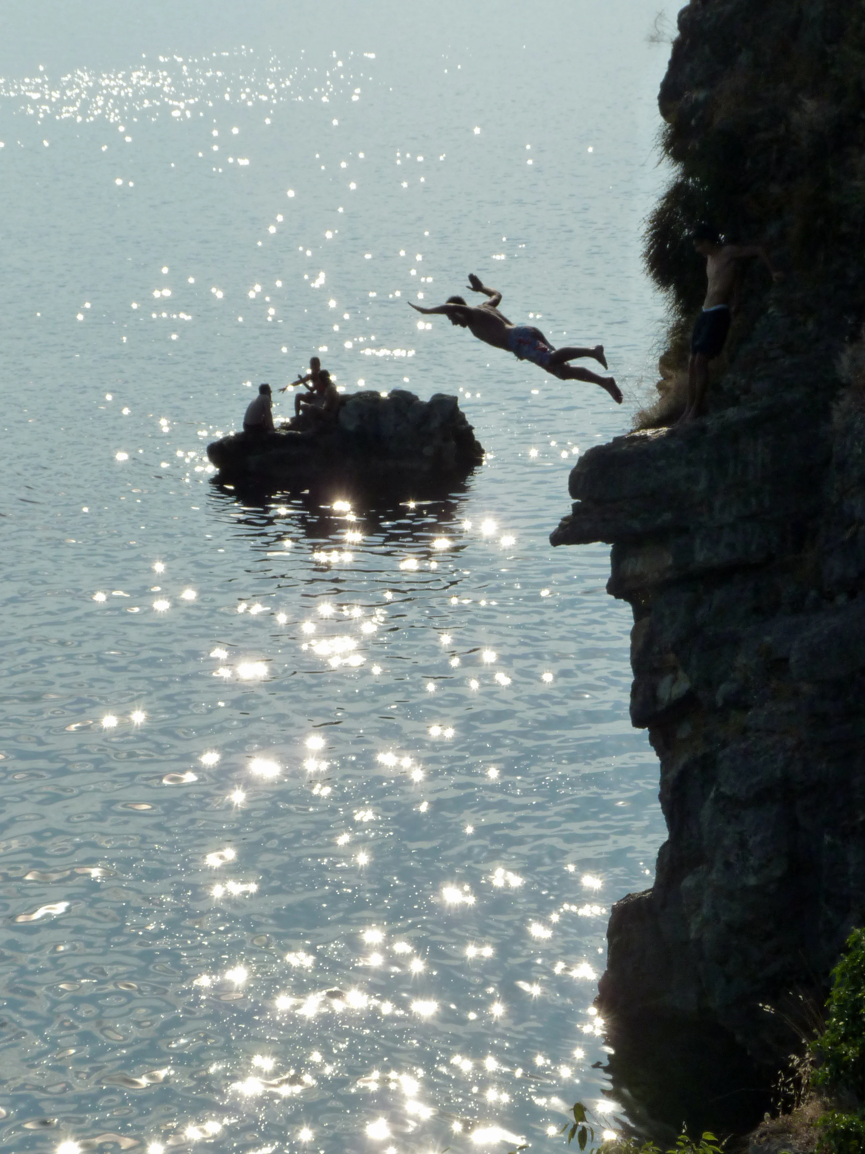 A person mid-air in a diving pose, jumping from a rocky cliff into the sparkling water of Lake Ohrid, with others visible on a nearby rock.