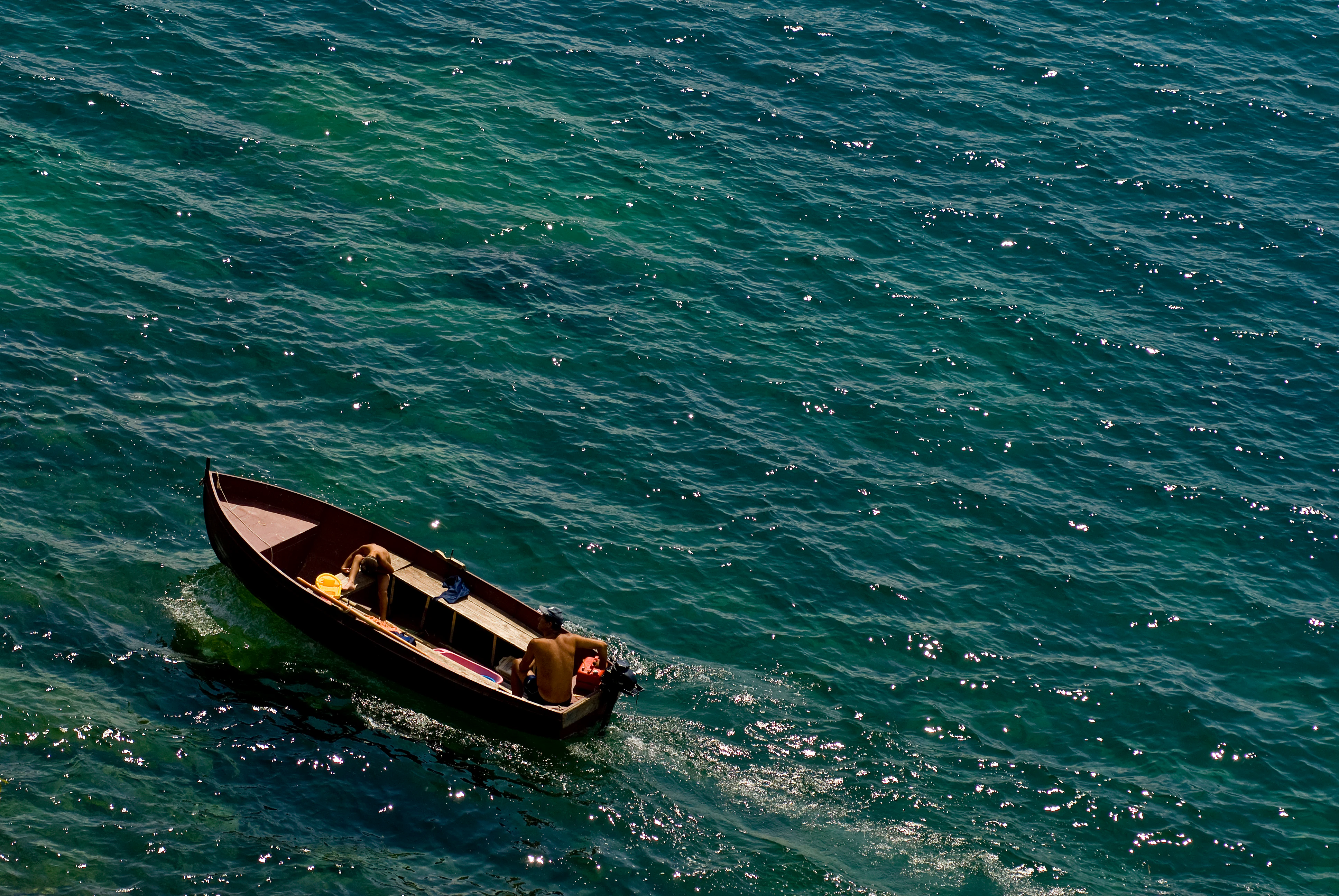 A high-angle view of a person operating a small motor boat moving across deep blue, sunlit lake water.