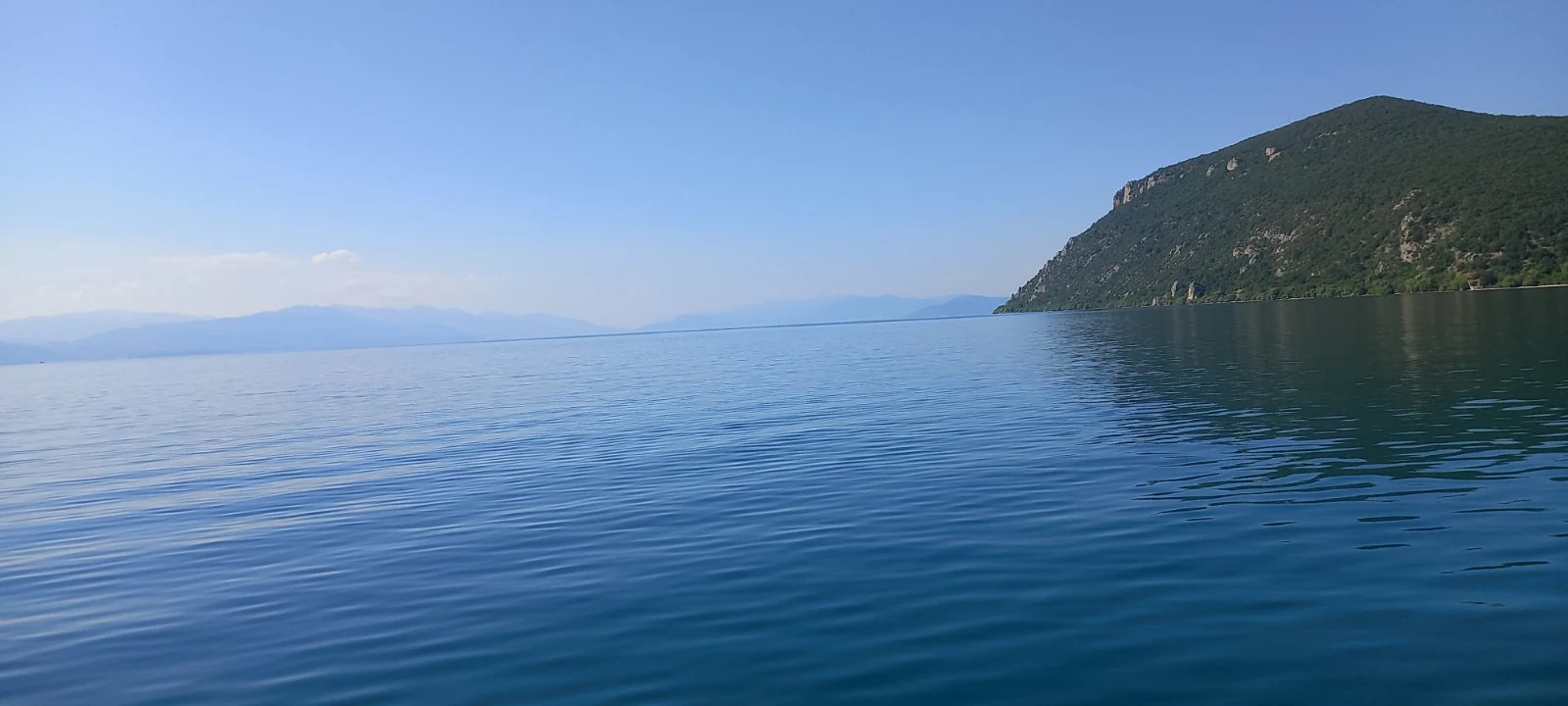 A expansive view of Lake Ohrid with deep blue water in the foreground and a green, sloping mountain rising from the shoreline on the right.