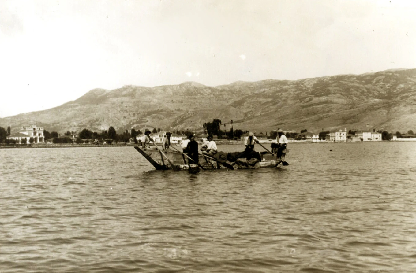 A black and white archival photograph showing several fishermen in a small, traditional wooden boat on Lake Ohrid, with low hills and distant shoreline buildings in the background.