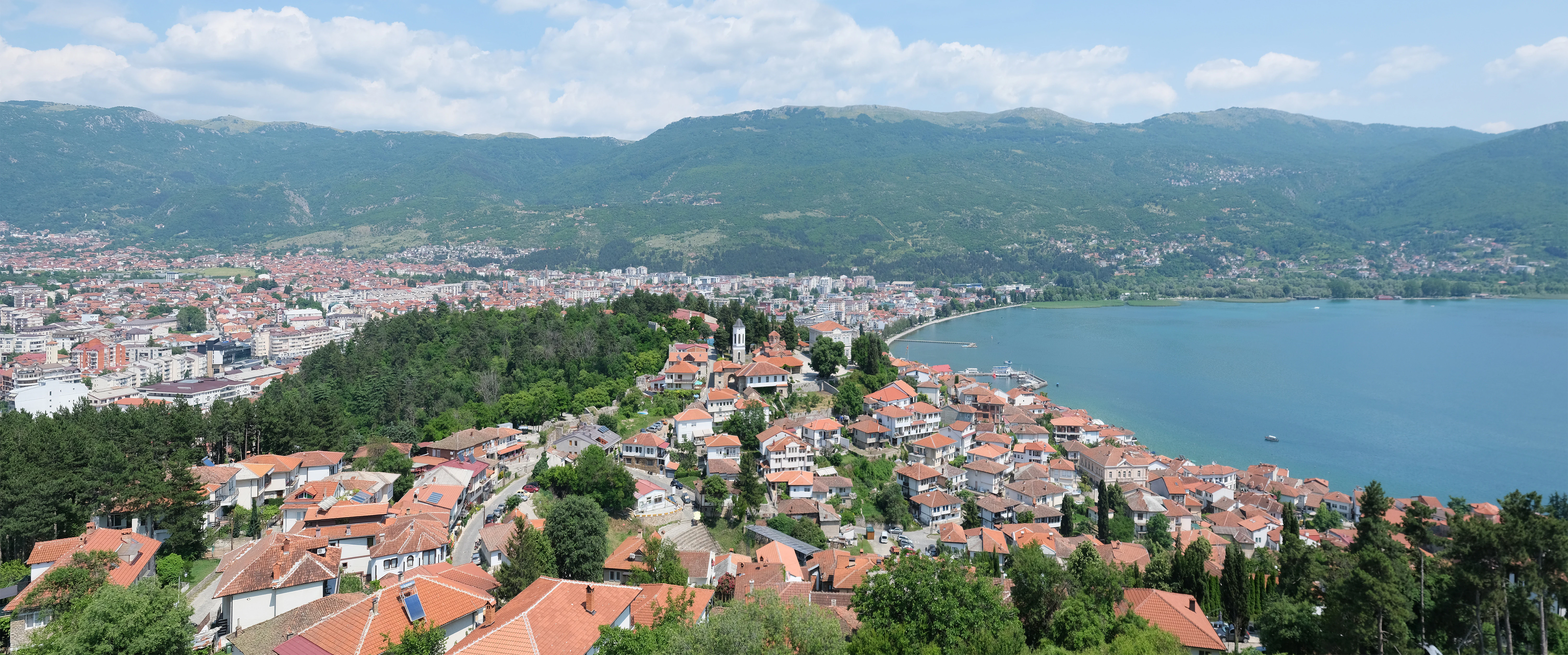 High angle panoramic view overlooking the red tiled rooftops of Ohrid city, the forested hillside, and the blue expanse of Lake Ohrid under a partly cloudy sky.