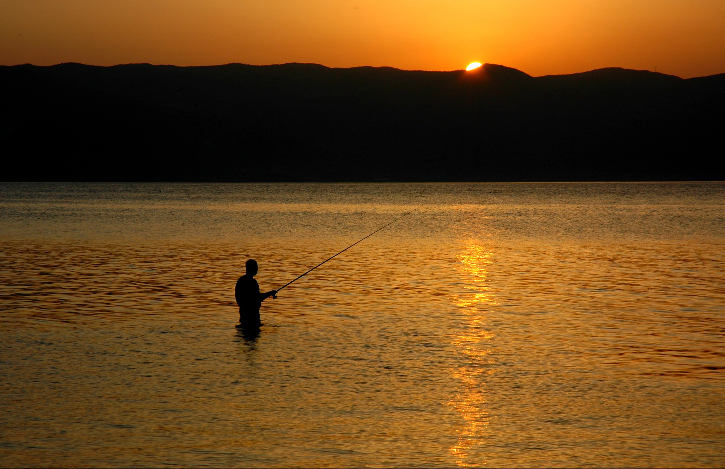 A silhouette of a person fishing with a rod while standing in the shallow water of a lake, with the sun setting behind distant dark hills.