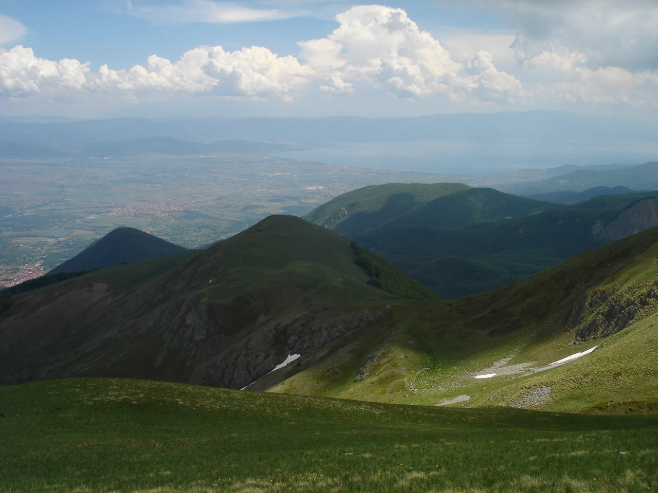 A panoramic high altitude view looking over green mountainous slopes and ridges towards a distant hazy lake.