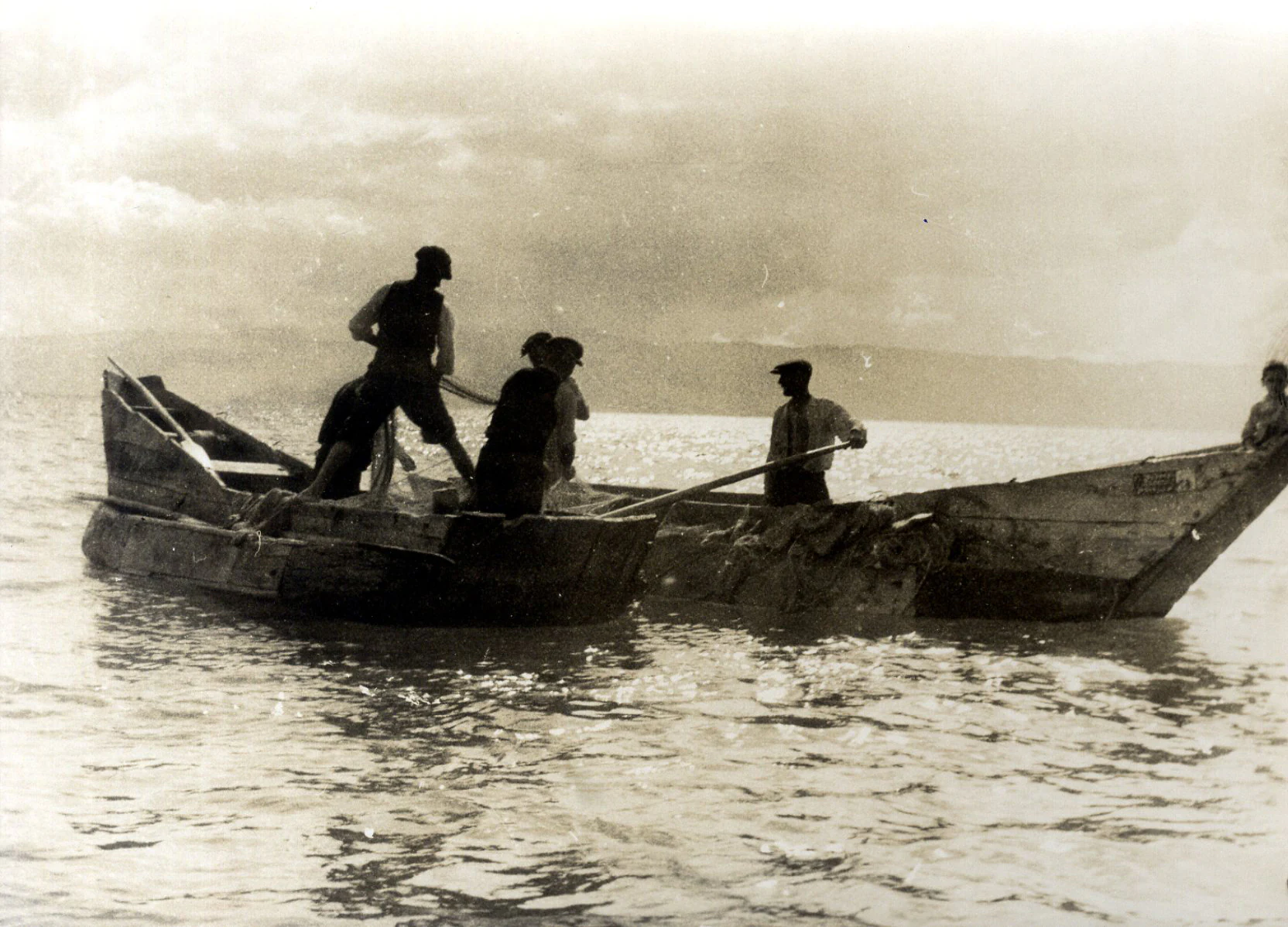 A sepia-toned archival photograph showing men in two small wooden boats on calm water, using oars and nets to fish.