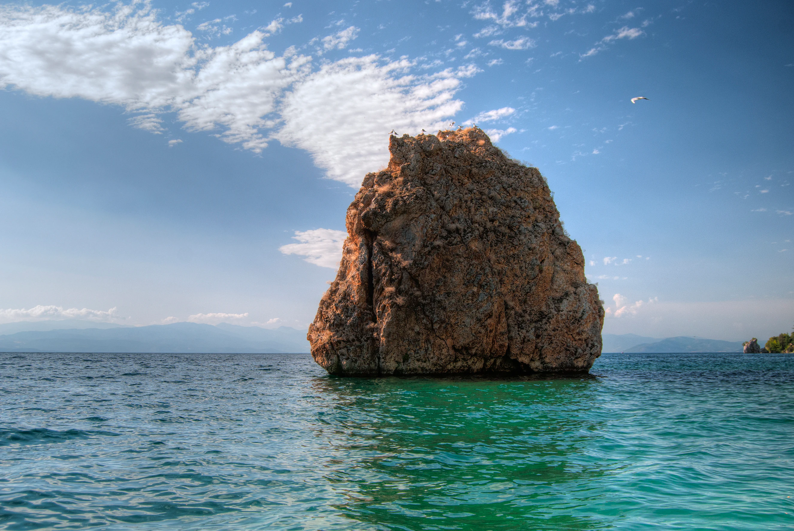 A large, weathered rock formation stands in the middle of clear turquoise lake water under a blue sky with scattered clouds.