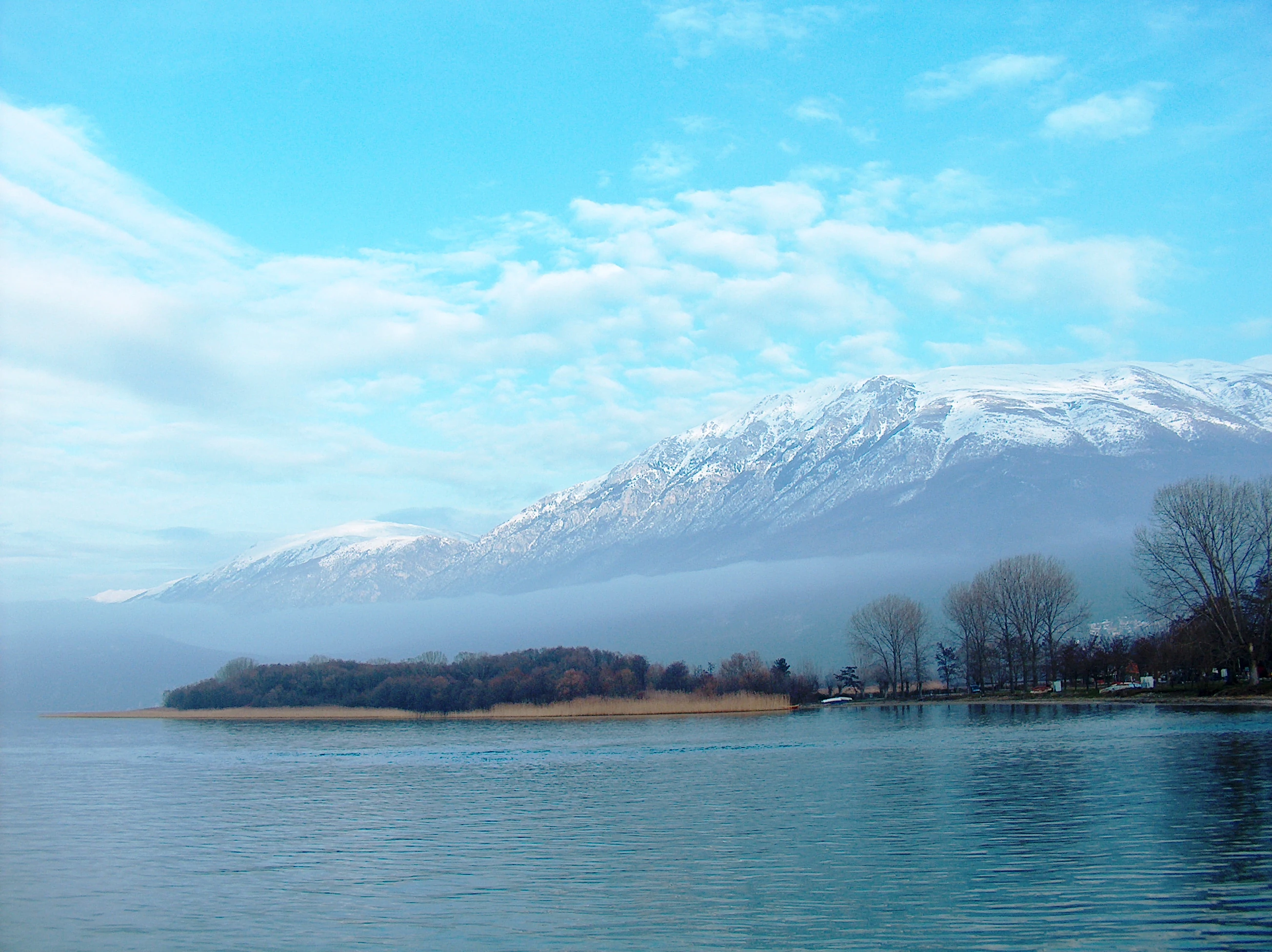 A wide shot of a calm blue lake under a light blue sky with wispy clouds, featuring a low-lying forested peninsula in the midground and a large, snow-dusted mountain range partially obscured by mist in the background.
