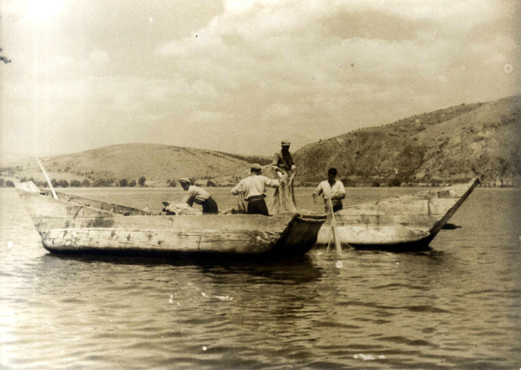 Black and white historical photograph showing several men working with fishing nets from wooden boats on Lake Ohrid with hills in the background.