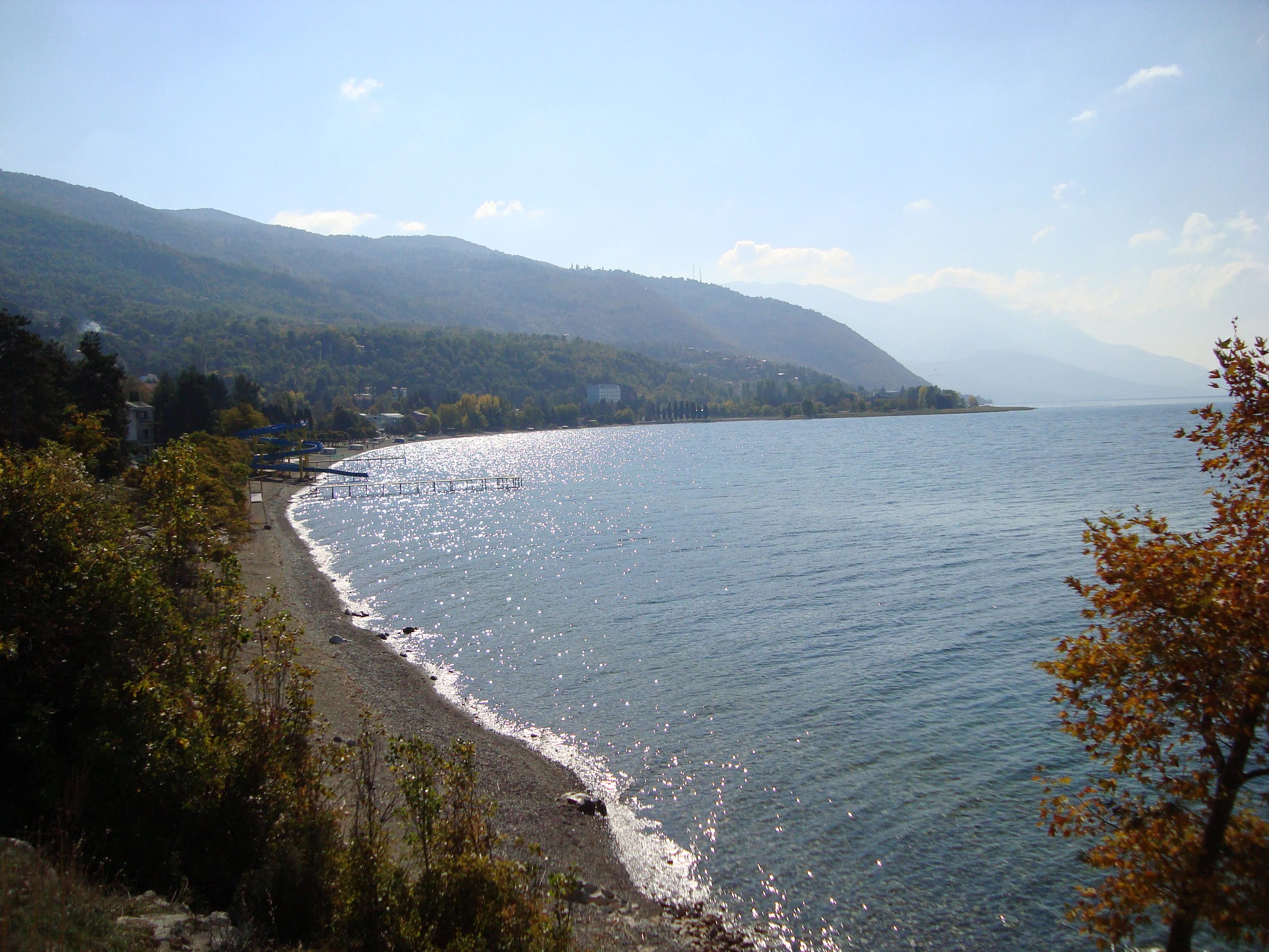 A wide angle view of the Lake Ohrid shoreline, with a narrow pebble beach on the left, calm blue water on the right, and tree-covered mountains sloping into the lake in the background.