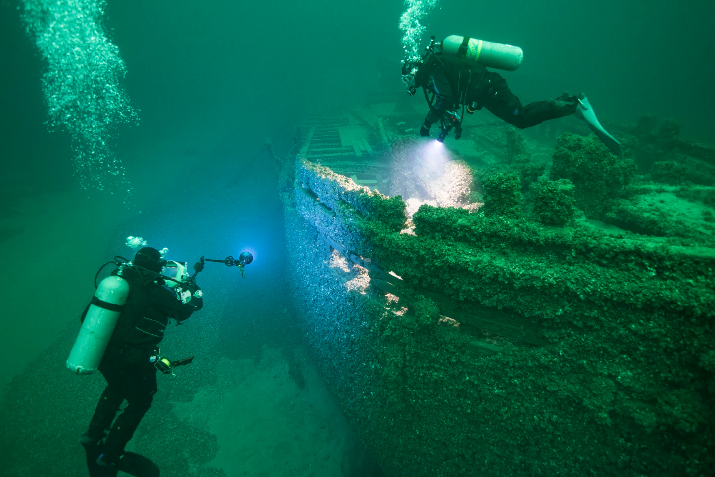 Two scuba divers in full gear exploring the wooden hull of a historic shipwreck underwater, illuminated by their dive lights.