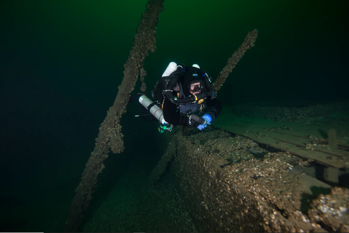 A scuba diver in full gear examines the submerged wooden wreckage of a ship in dark, deep water.