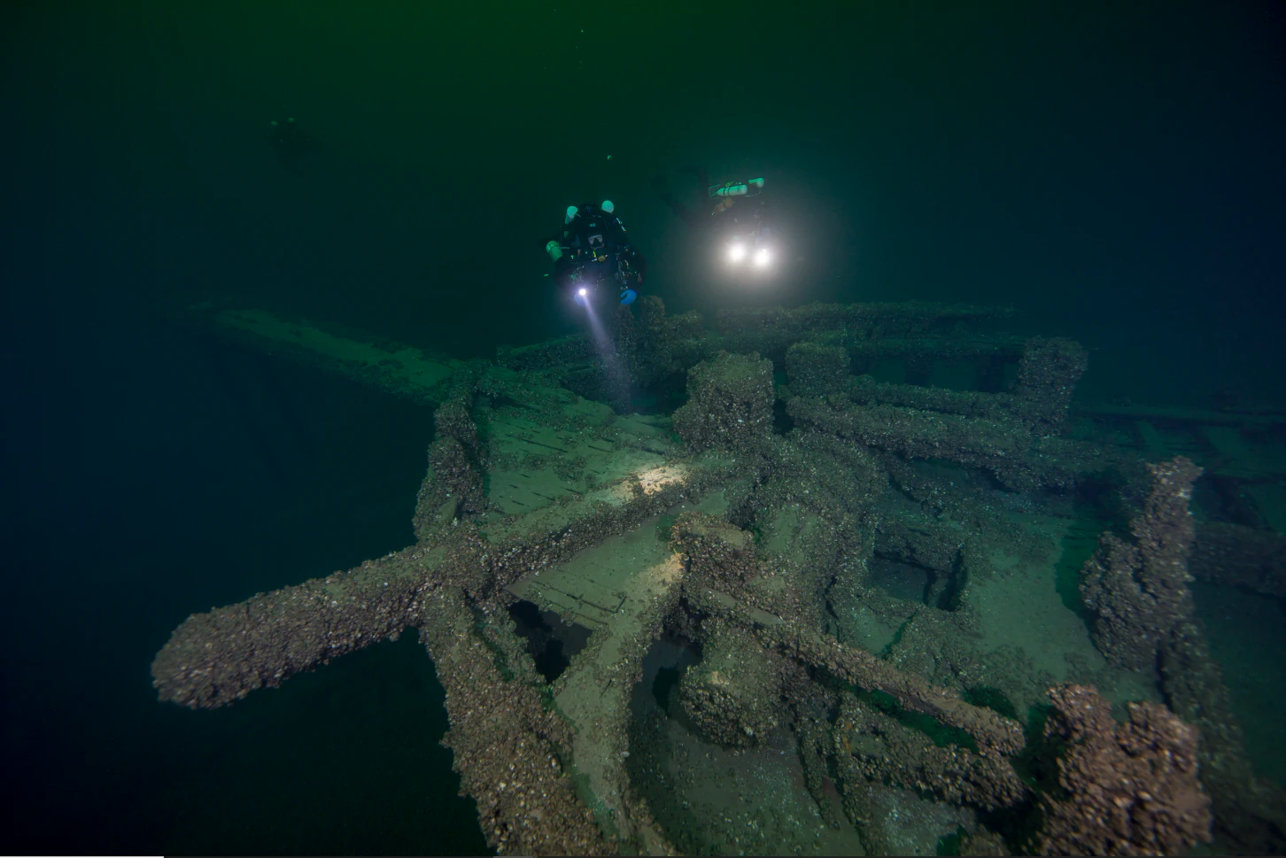 Underwater view of two scuba divers using flashlights to explore the wooden remains of a sunken shipwreck on the lake bed.