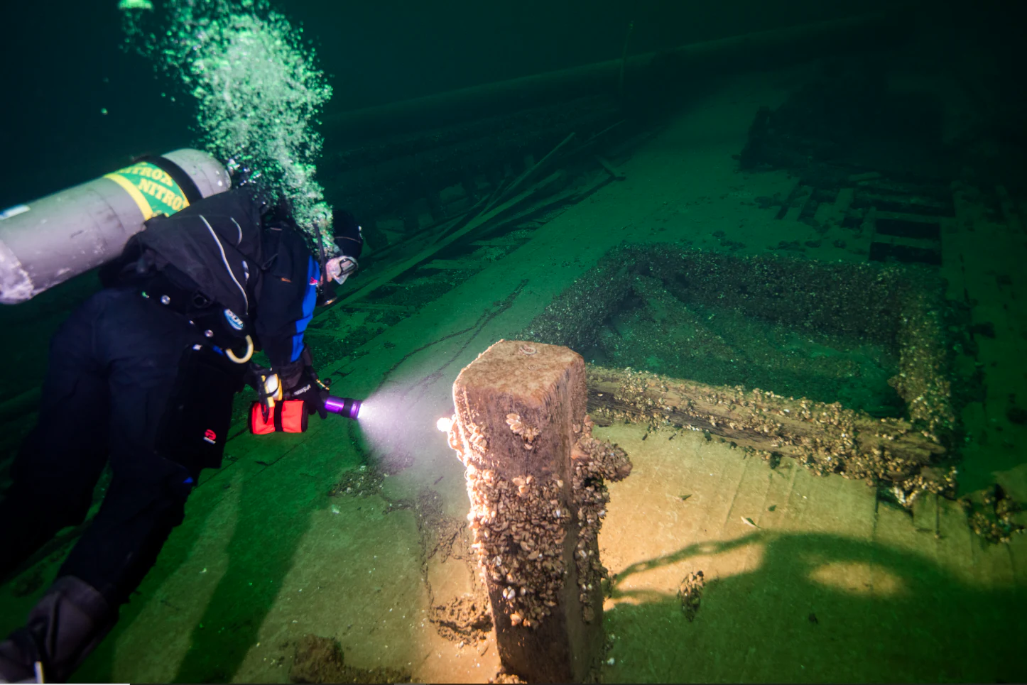 An underwater view of a scuba diver in dark gear using a flashlight to explore the wooden wreckage of a submerged ship.