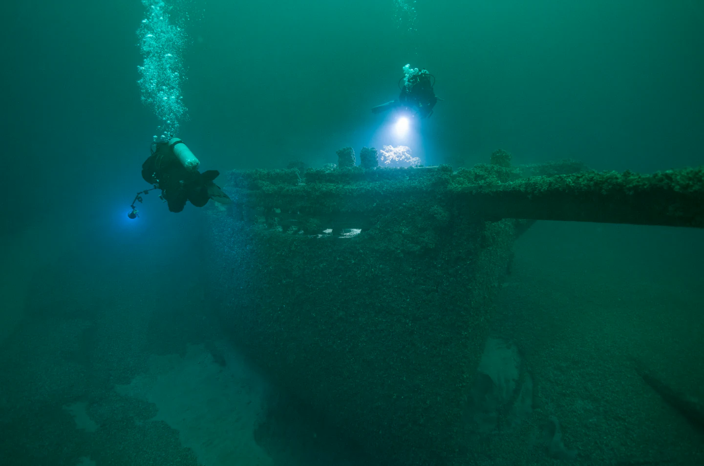 Underwater view of two scuba divers using flashlights to inspect the wooden wreckage of a historic schooner on the lakebed.