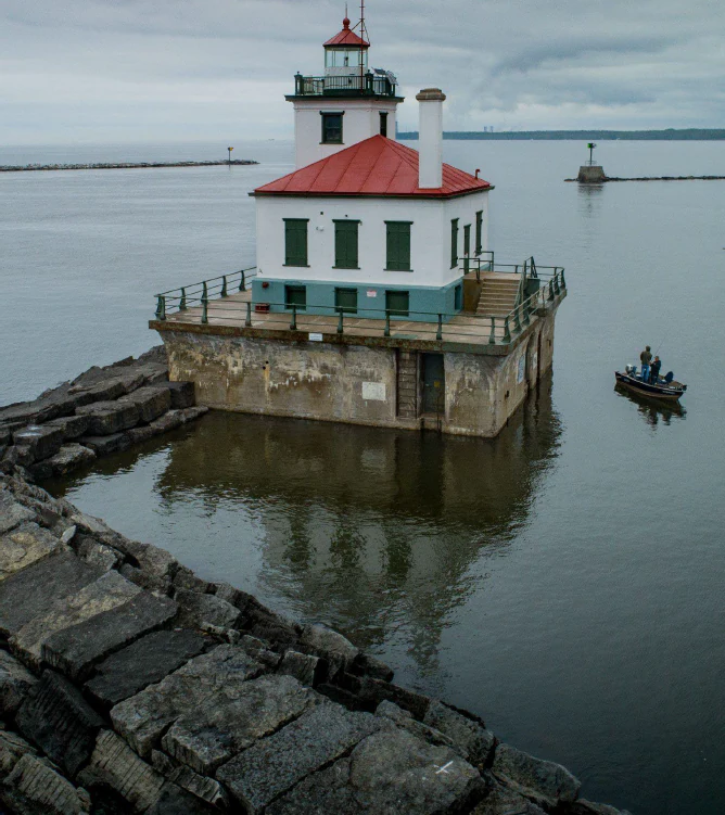 A white square lighthouse with a red roof and green trim, built on a stone-faced concrete pier in calm water, with a small boat nearby and a rocky breakwater in the foreground.
