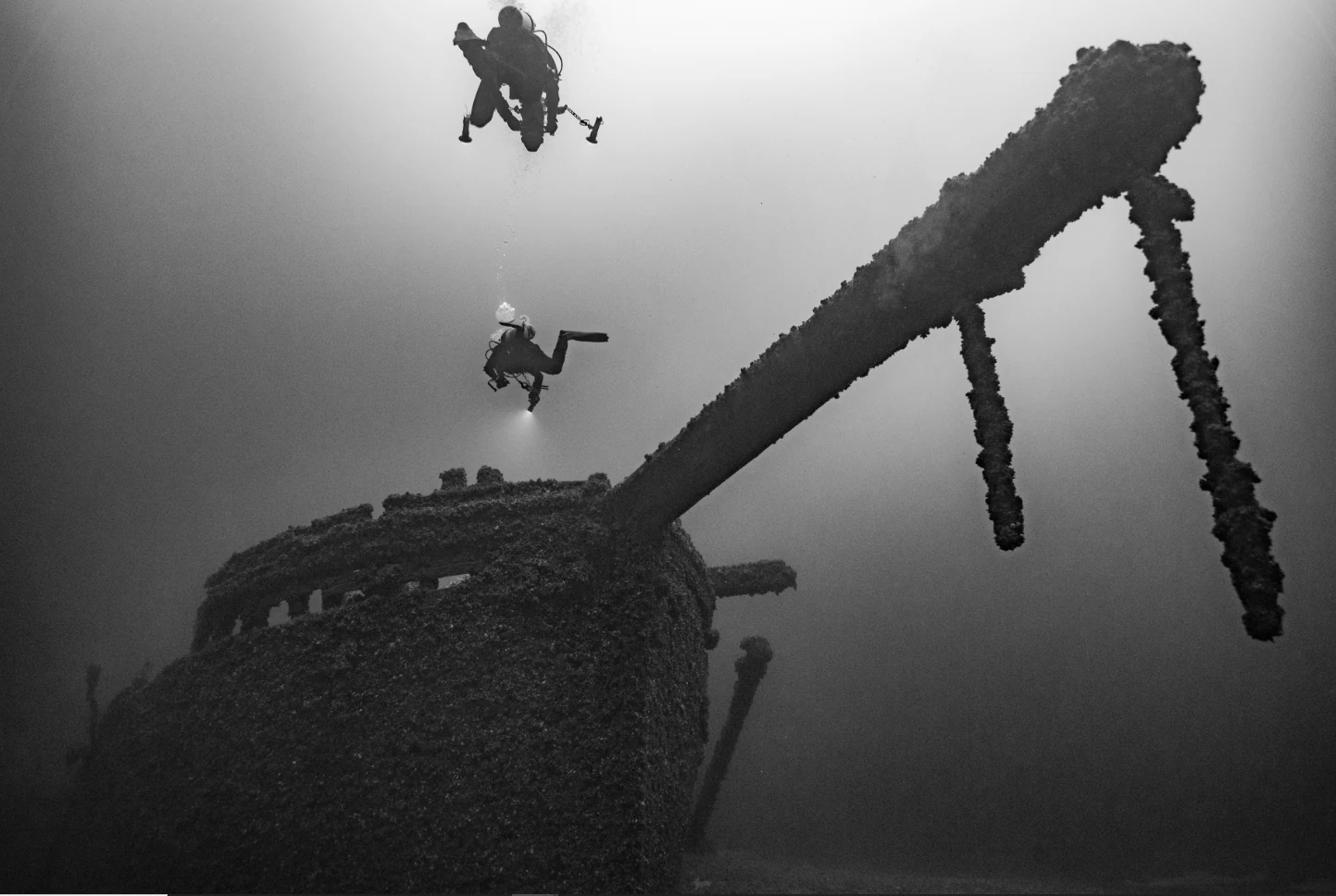Two scuba divers swimming near the prominent bowsprit and hull of an underwater shipwreck, rendered in high-contrast black and white.