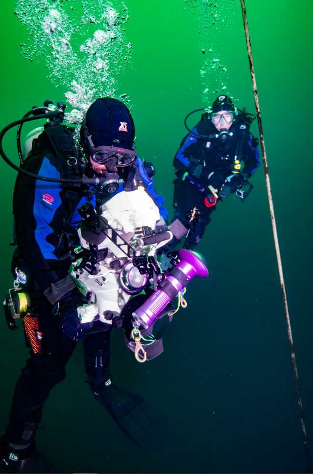 Two scuba divers suspended in deep green water while using a professional underwater camera setup to film a historic shipwreck site.