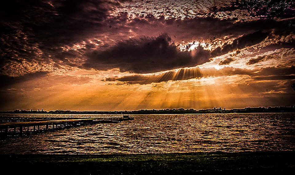 Lake Palić at sunset with dramatic clouds and sun rays breaking through, showing a wooden dock on the left side and calm water reflecting the sky