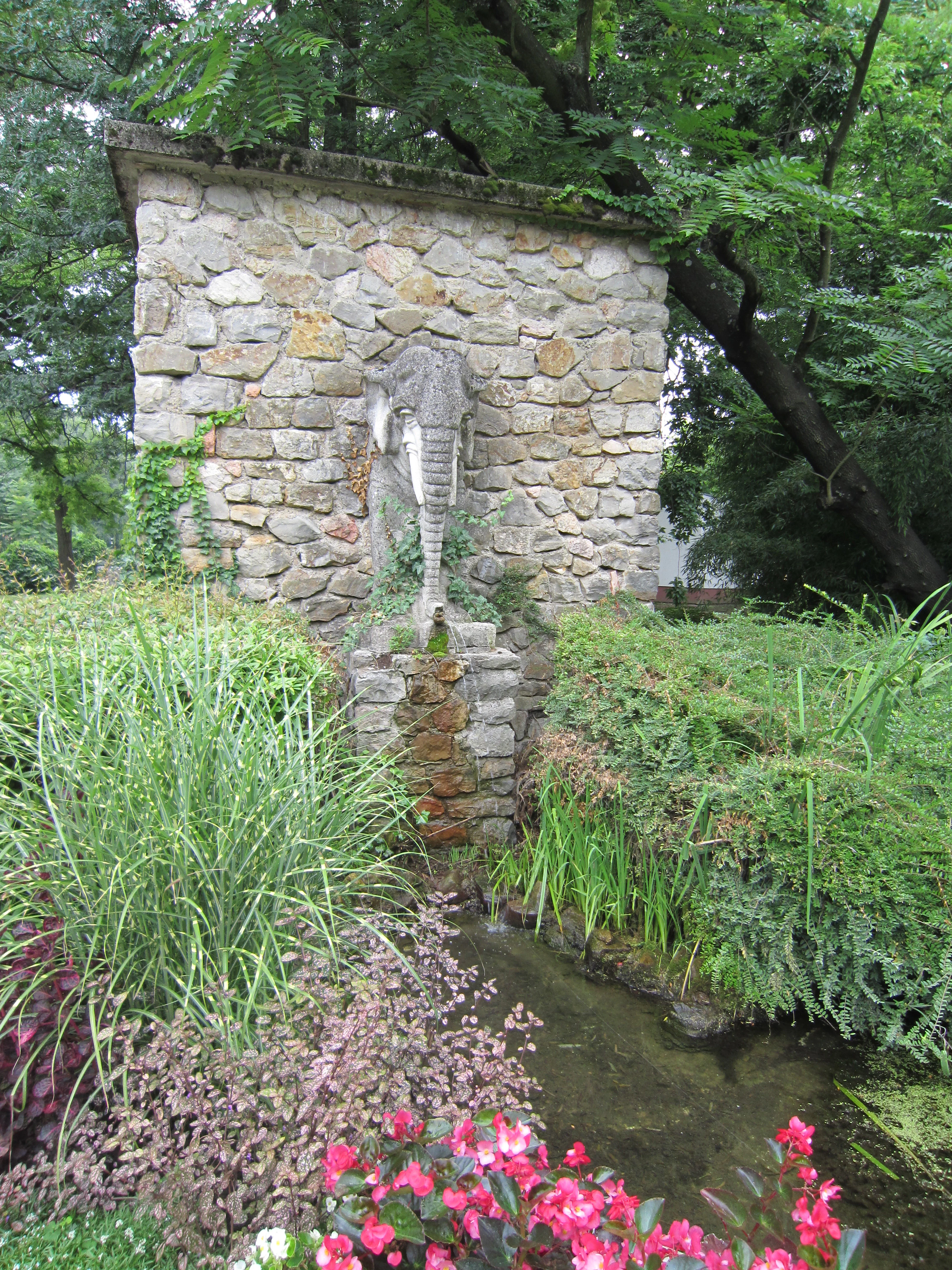Stone wall with elephant statue fountain, surrounded by green plants and pink flowers with a small stream
