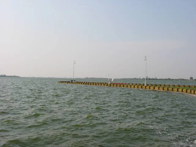 A wide view of Lake Palić showing choppy water with a long yellow and brown pier structure extending from the right side toward the horizon under a hazy sky