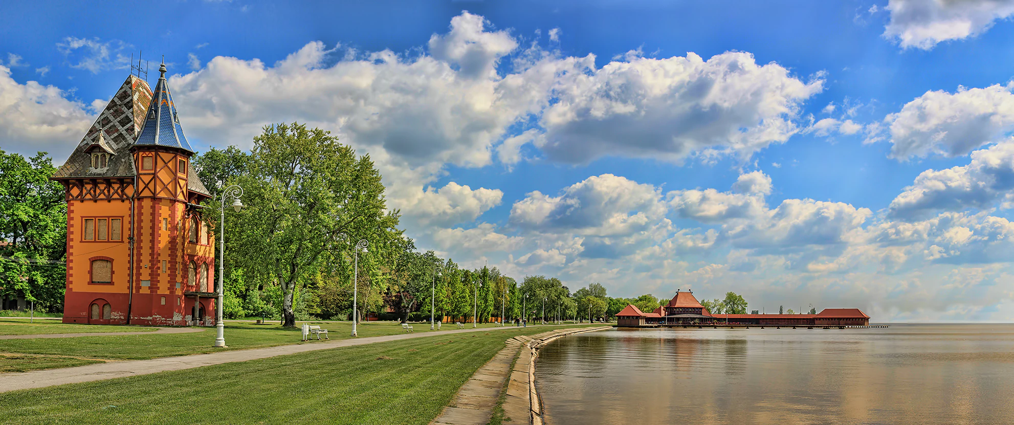 Panoramic view of Lake Palić showing a red-brick building with a steep roof, paved pathway, green grass, trees, and calm lake waters under a partly cloudy sky.