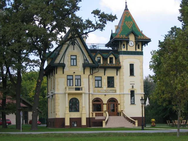 Two-story yellow building with green trim and clock tower surrounded by trees and grass
