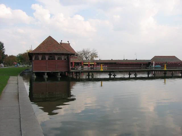Wooden building on stilts over calm lake water with a long pier and grassy shoreline