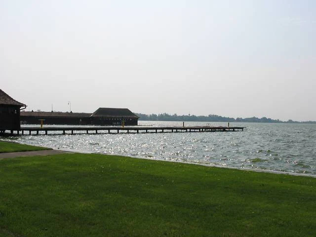 A large body of water with a wooden pier extending from a grassy shore, featuring buildings on the pier under an overcast sky