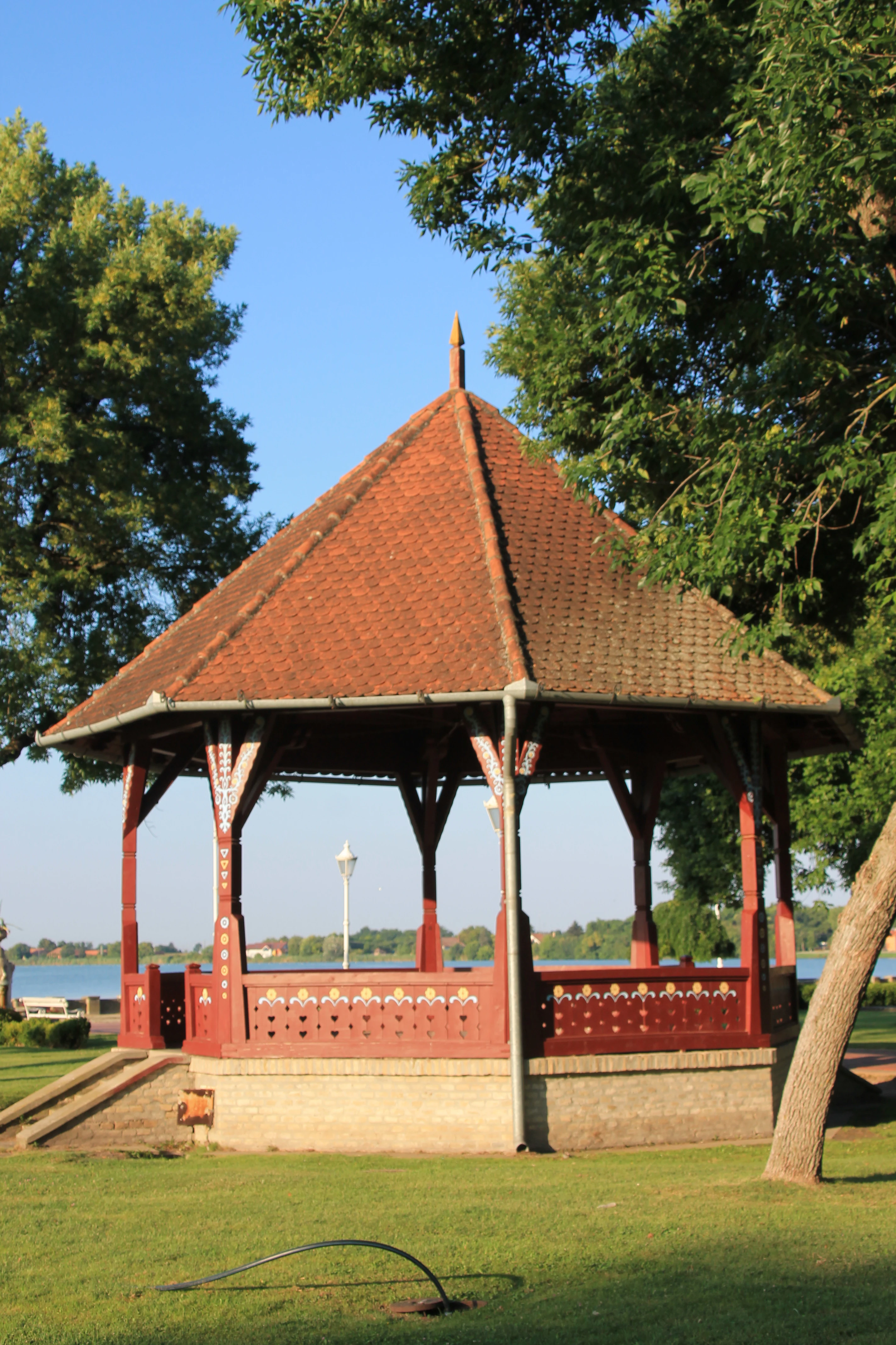 Red-tiled wooden pavilion with decorative railings on a grassy area, surrounded by trees and a body of water in the background