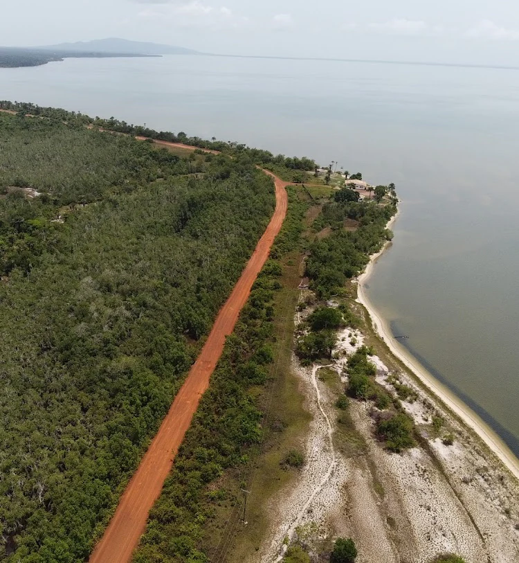 An aerial view captures a red dirt road running parallel to the sandy shoreline of a large, calm lake, with dense green forest on one side.