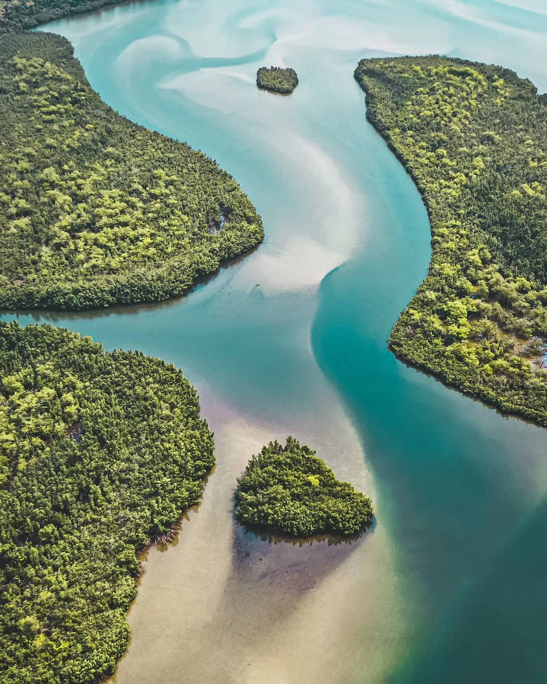 An aerial view looking down at a winding turquoise waterway surrounded by dense green forest, featuring small forested islands within the shallow sandy channels.